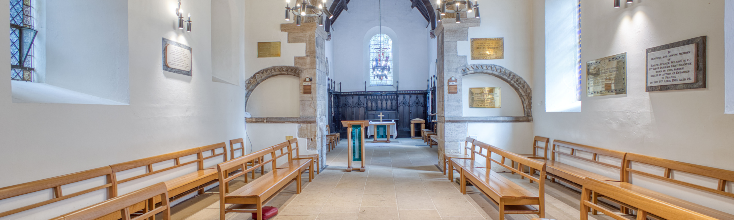 An empty, brightly lit historic chapel with rows of wooden pews lining both sides of a central stone aisle leading to the altar.