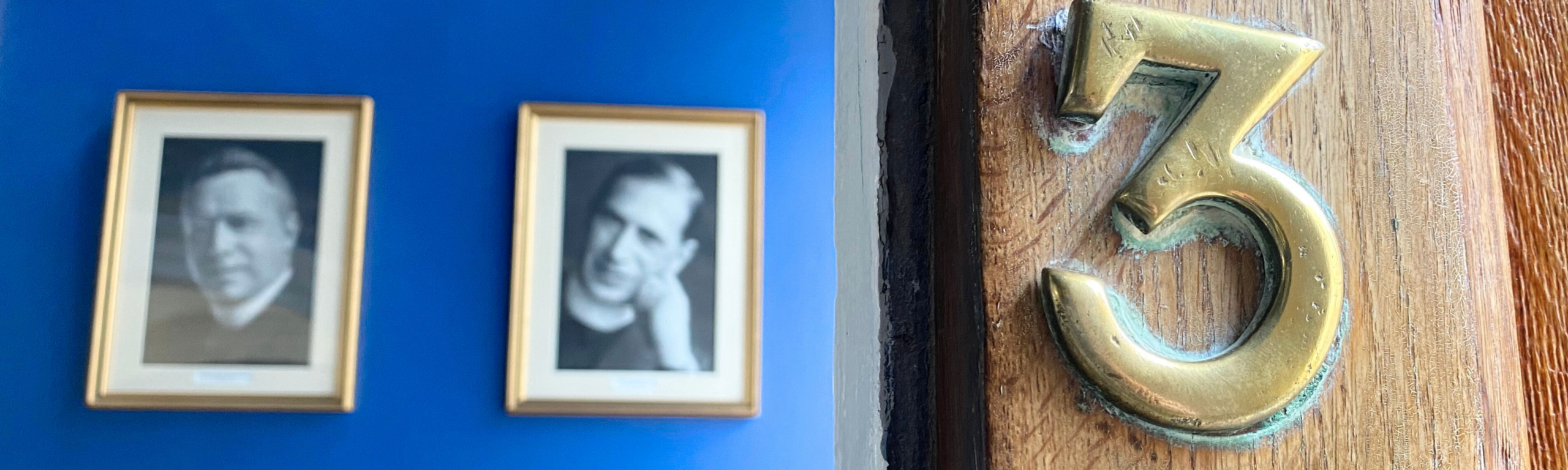 The brass number three on the St John's College main entrance door, showing a glimpse of historic alumni portraits on a blue wall inside