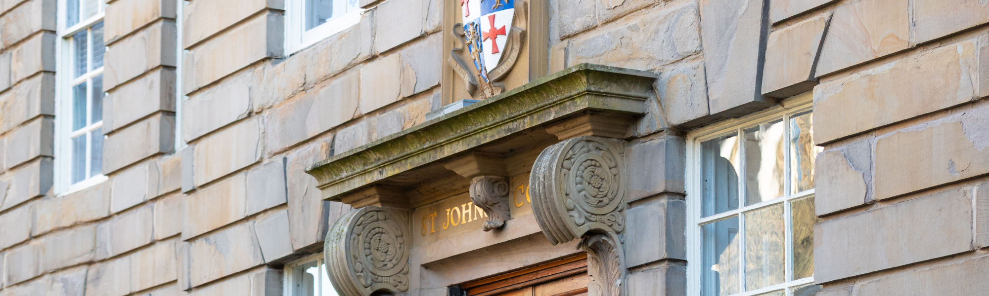The grand stone entrance of St John's College, featuring an ornate carved lintel with the college name engraved in gold, topped by the vibrant official coat of arms