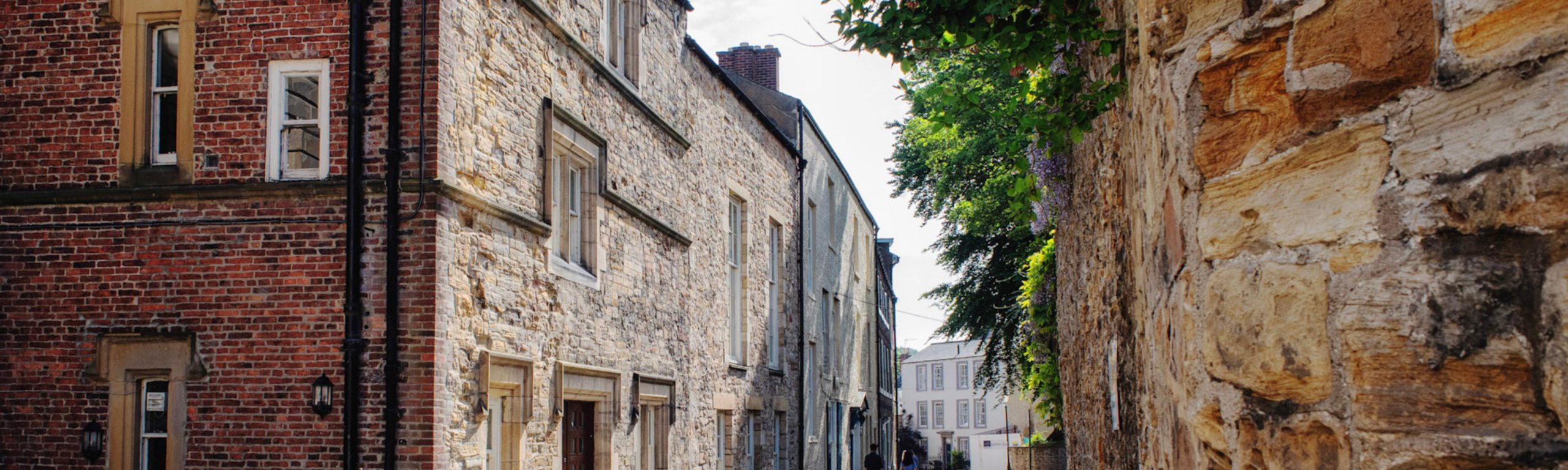 A sunny view looking down a narrow historic street lined with traditional red brick and stone college buildings, with lush greenery overhanging a wall on the right.