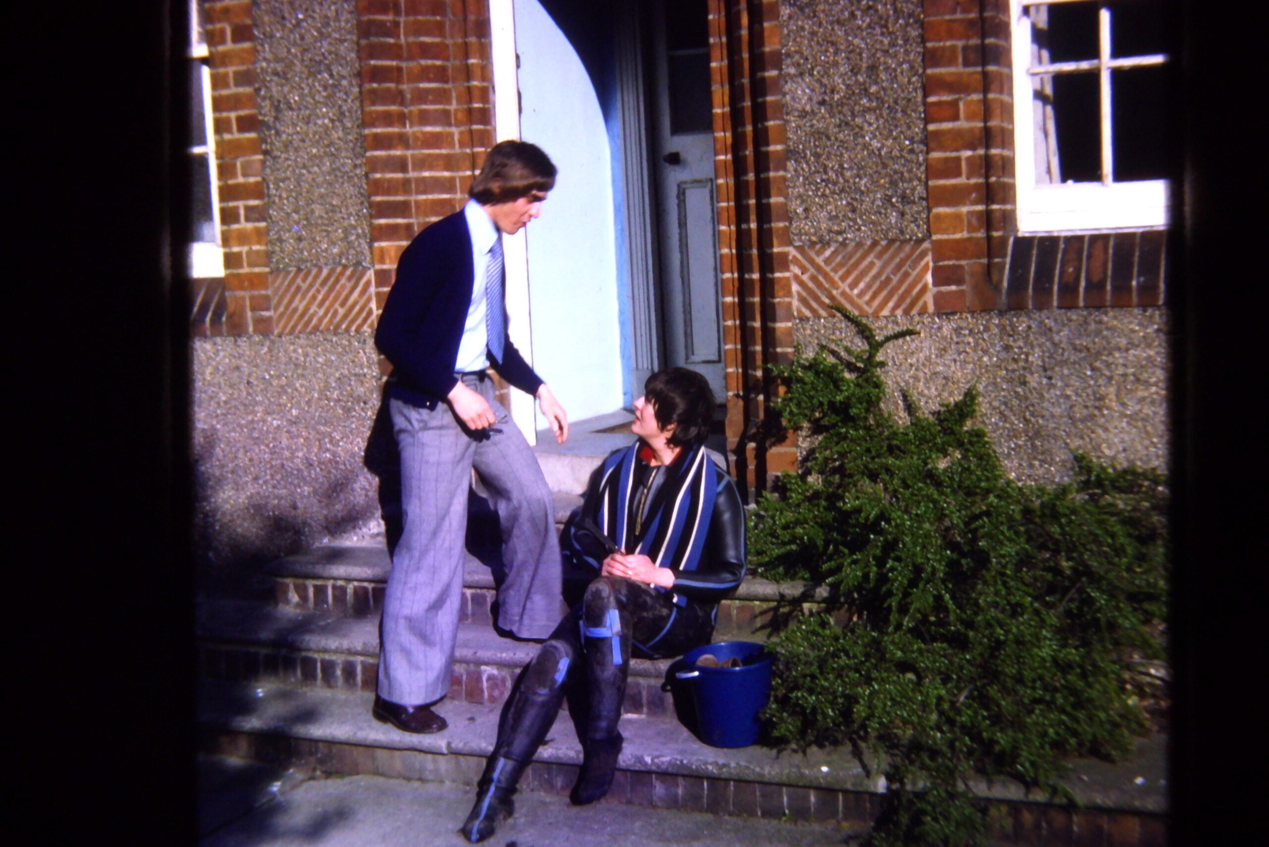 A young man stands to the left of a young woman sitting on the steps of a brick building