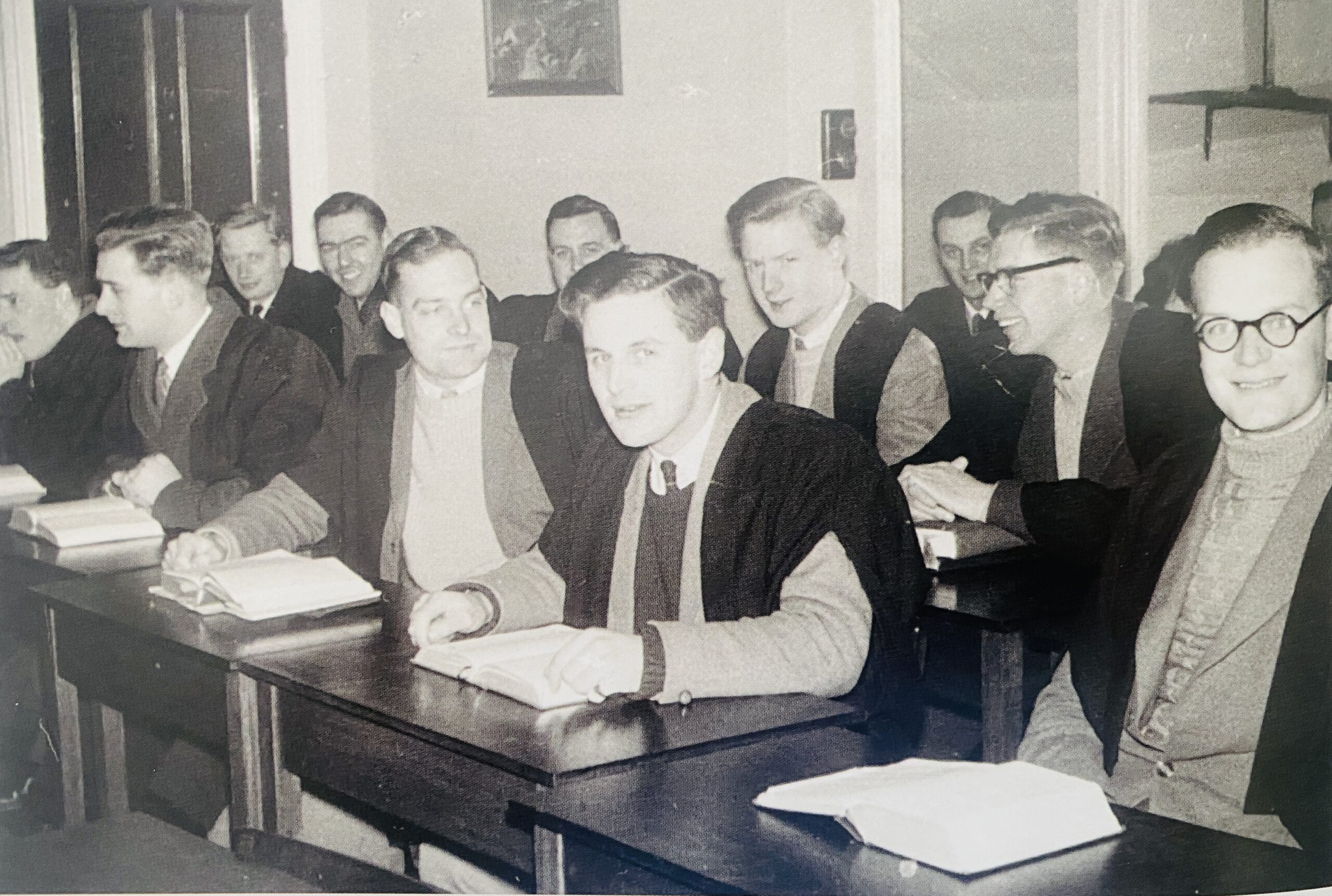 black and white photo of young men in academic dress sit a desks packed closely together