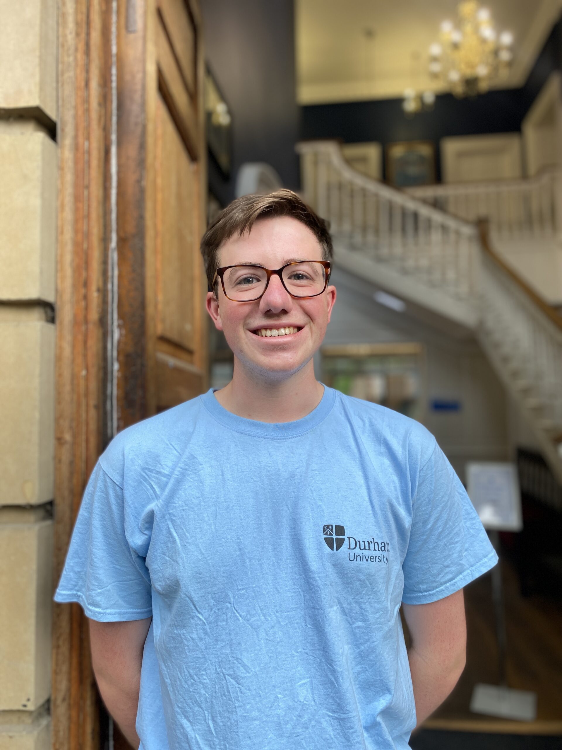 A young man in glasses and a light blue t shirt smiles at the camera from an open door way